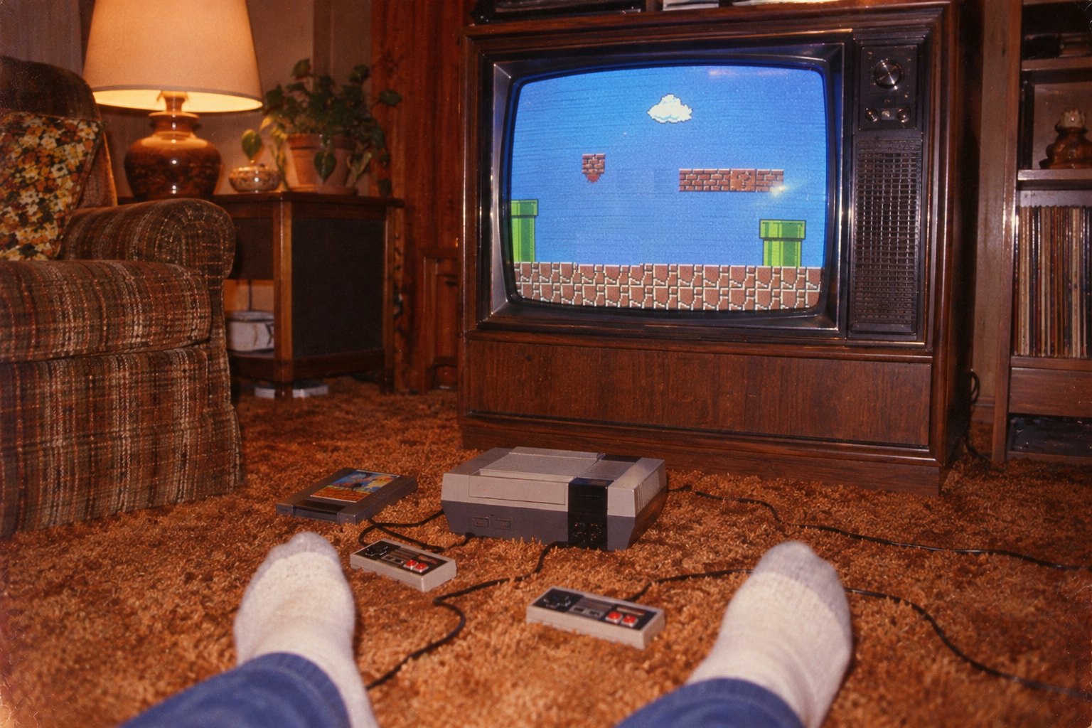 A 1985 living room from the floor: socked feet on a brown shag carpet, a grey console and two rectangular controllers in front of a wood-cabinet television showing a side-scrolling platform game, a plaid armchair and a lamp beside it