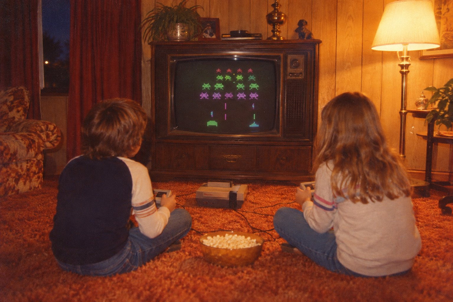 Two kids on an orange shag carpet in front of a wood-cabinet television playing an invaders game, a bowl of popcorn between them, a lamp glowing beside the TV